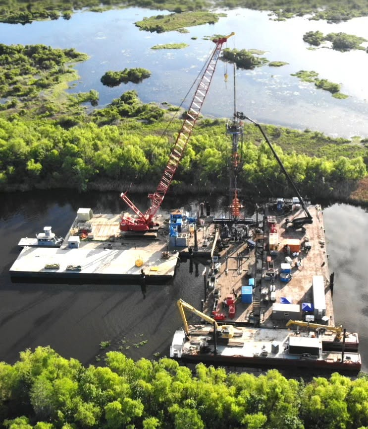 Aerial view of a barge-mounted drilling rig operating in a wetland, surrounded by trees and shallow water with patches of vegetation.