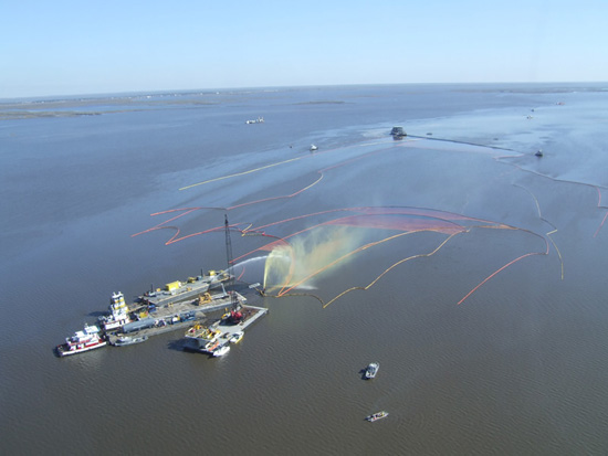Aerial view of an oil spill on coastal water, with response vessels, orange containment boom, and a visible oil sheen near a marshy shoreline.